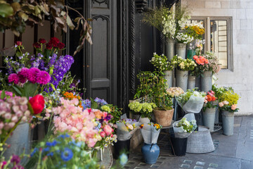 A diverse and colorful array of flowers are presented in various metal buckets and pots outside a shop.