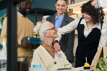 Senior Caucasian woman sitting at desk wearing retired badge smiling at multiethnic group of young...