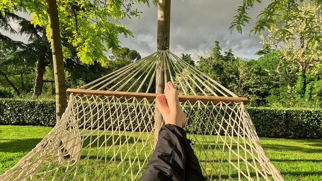 A persons legs in pants and bare feet relaxing in a hammock strung between trees in a green garden, concept of relaxation, leisure, outdoor comfort, POV