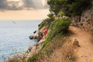 A woman in a red towel returns from a wild, rocky beach, climbing the scenic coastal path in Saint-Jean-Cap-Ferrat on the French Riviera.