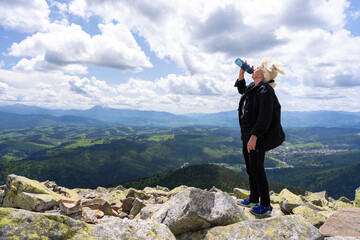A woman is drinking water while standing on top of a mountain.