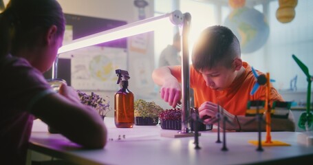 Smart Young Boy and Girl Sitting at Table, Growing Experimental Microgreens Under Grow Light, Using...