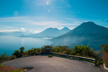 Scenic view of a lush forest and calm lake in Guatemala, with a green volcanic hill rising near the shoreline and vast mountains fading into the misty background under soft clouds.