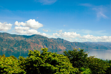 Scenic view of a lush forest and calm lake in Guatemala, with a green volcanic hill rising near the shoreline and vast mountains fading into the misty background under soft clouds.