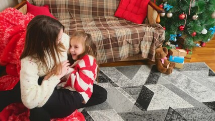 Happy family having fun at home for Christmas. Mother tickles her daughter sitting on the floor. Festive cozy interior - Powered by Adobe