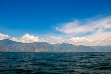 Scenic view of a lush forest and calm lake in Guatemala, with a green volcanic hill rising near the shoreline and vast mountains fading into the misty background under soft clouds.