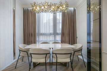 The interior of the living room with an elegant design. White room decor. Dining table with comfortable beige chairs. Beautiful ceiling lamp.