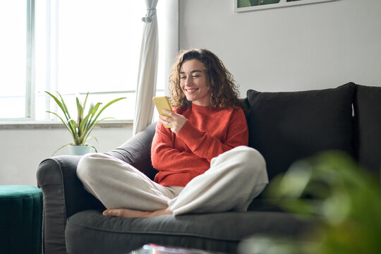 Smiling cheerful lady laughing holding smartphone having fun while buying in ecommerce shop or watching funny videos online. Young happy woman using mobile cell phone sitting on couch at home. - Powered by Adobe
