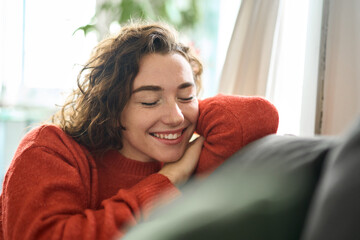 Naklejka na ściany i meble Young smiling pretty curly woman relaxing sitting on couch at home and chilling. Happy relaxed calm beautiful lady enjoying peace of mind and comfort of soft new sofa with eyes closed. Close up.