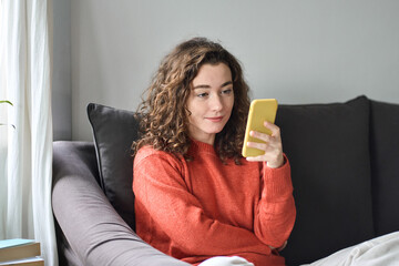 Pretty young woman sitting on sofa using cell phone, holding smartphone, looking at cellphone doing online shopping choosing fashion clothes in mobile apps, checking social media commercials.