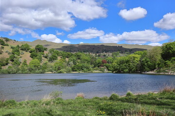 Panorama of Rydal Water surrounded by green hills and trees under a partly cloudy blue sky in the Lake District, England, United Kingdom.