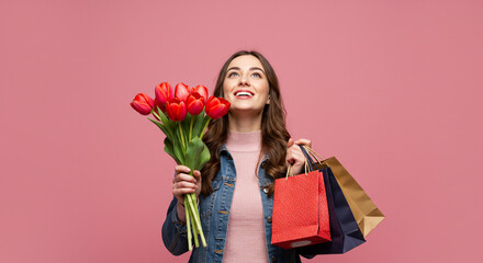 Woman holding red tulips and shopping bags smiling looking up against a pink background in a studio shot