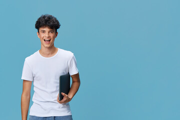excited young man holding a laptop standing against blue background, wearing casual white t-shirt...