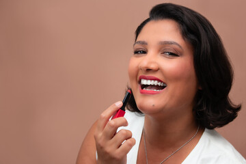 Beautiful brown skin woman laughing and applying red lipstick. Perfect for cosmetics, make-up, beauty, self-care, self-love. Ample copy space on beige background.