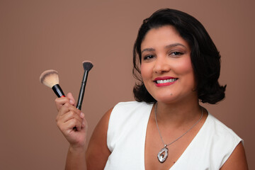 Friendly woman in white tank top smiling and holding make-up brushes. Suitable for beauty, cosmetics, makeup and embellishment themes. Horizontal studio portrait with copy space on beige background.