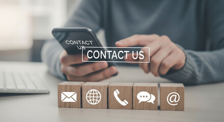 Person using phone with contact us message above wooden blocks with contact icons on the table top