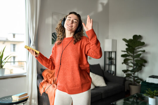 Candid photo of happy pretty young woman wearing headphones dancing at home. Cheerful girl listening music on mobile phone, singing song feeling relaxed standing in modern living room interior.