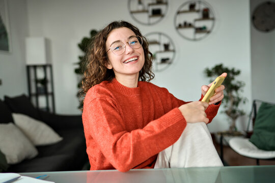 Young happy woman holding smartphone using cellphone modern technology, looking at camera with mobile, checking cell phone app, texting typing messages, browsing internet for shopping sitting at home. - Powered by Adobe
