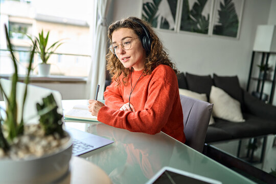 Busy young woman student wearing headphones using laptop elearning or remote working at home looking at computer watching webinar, learning online seminar or video calling for work meeting.