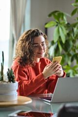Happy young lady using smartphone social media apps at home. Smiling woman holding cellphone modern ai applications, looking at mobile, checking cell phone, texting chatting sitting at table, vertical