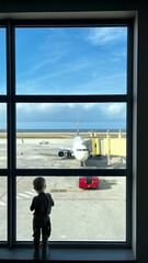 Silhouette of little blond boy is looking out of big window in airport, watching transport circulation and vehicle movement around aircrafts. Toddler enjoys traveling with airplane and vacation