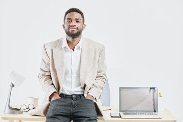 Confident young man with beard wearing beige blazer and white shirt standing in modern office with...