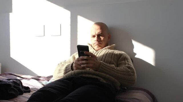 49 year old Brazilian man sitting on the bed feeling sad after reading news on his smartphone.