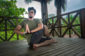 Young man doing a yoga asana position in a forest wooden building with plants and green trees. Stretching exercise for good health, balance, positive mind, mental health and spiritual equilibrium