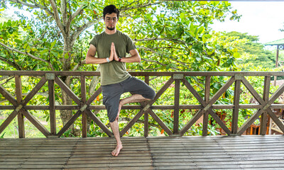 Young man doing a yoga asana position in a forest wooden building with plants and green trees....