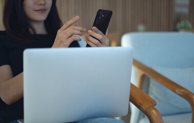 Asian woman using mobile phone, working on laptop computer on her lap at home. Casual business...