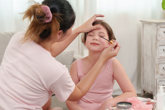 Asian single mother applying eye shadow on daughter's eyelids, daughter smiling with closed eyes during beauty learning time, joyful family bonding, maternal love, trust and encouragement in cozy home - Powered by Adobe