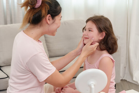 Asian single mother cupping daughter's face affectionately during happy beauty session, smiling joyfully, nurturing connection, trust and love flowing between parent and child, home setting shared
