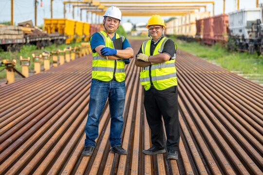 Team Work Asian: Two Professional Engineers with Arms Crossed at Railway Site Outdoor Construction: Smiling Workers in Safety Vests at Railroad Excellent Team: Engineers in Hard Hats.