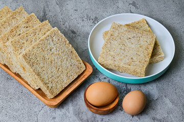 Close up view of whole wheat sliced bread on plate and long wooden plate with two chicken eggs. grey or gray cement as background.