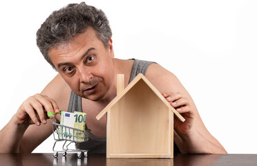 A poor middle-aged man in a sleeveless T-shirt, a miniature wooden house and a 100 euro note
