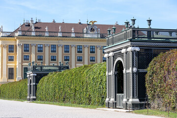 
A palace complex with a yellow Baroque building in the background, green, clipped hedge walls, and...