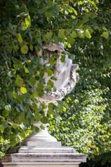 
a large, white vase with carved decorations, partially hidden behind green leaves and trees.