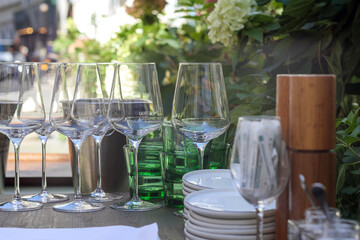 
A table setting with glasses, plates, and other dishes prepared for an outdoor event with greenery and flowers in the background.