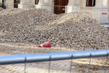 
A scene of a construction site with two large piles of gravel or rubble, a red cone lying in the...