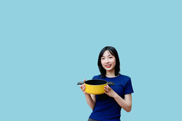 Young woman holding yellow cooking pot against a blue background while smiling in a modern kitchen...