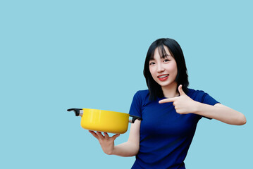 Asian woman presenting a yellow cooking pot against a solid blue background while smiling and...