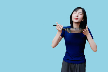 Young Asian woman enjoying delicious food from a pan against a blue background while expressing...