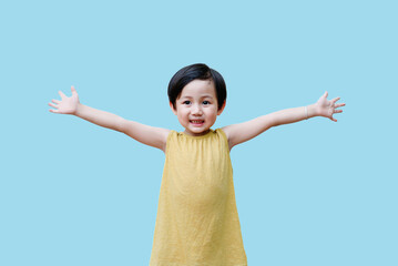 Child with short hair in a yellow dress joyfully spreading arms against a light blue background,...