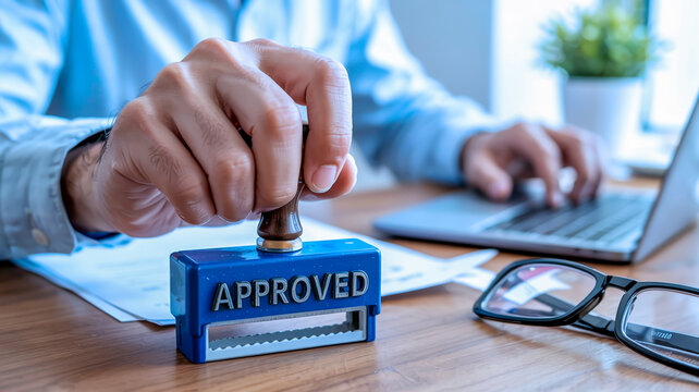 A close-up business photograph showing hands working at a wooden desk. In the foreground, a hand is holding a blue rubber stamp with a black handle