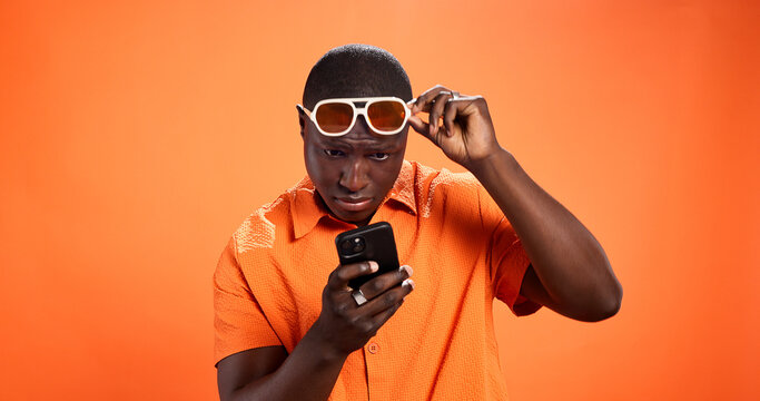 Black man, phone and shocked in studio with chat, thinking and reading post on orange background. African person, smartphone and doubt with fake news, gossip and confused on social media with choice