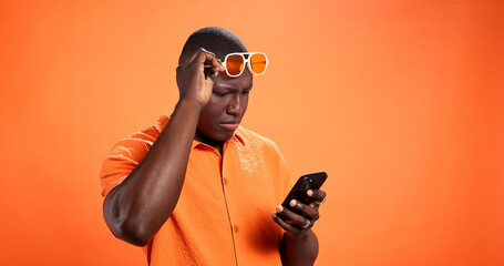 Fototapeta na wymiar Black man, phone and confused in studio with chat, thinking and reading post on orange background. African person, smartphone and doubt with fake news, gossip and scroll choice on social network
