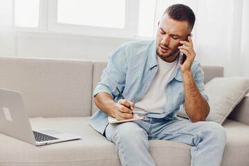 Fototapeta premium Focused man making notes while talking on the phone, dressed casually in a light denim jacket. The modern setting features ample natural light.
