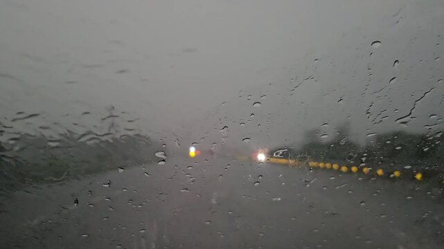 A close-up view through the rain-soaked west glass window of a motor vehicle reveals blurred lights, passing cars, and traffic reflections, as windshield wipers sweep across the glass falling rain.