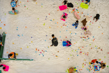 Top view of children playing on artificial snow with colorful toys and sleds in an indoor playground area, creating a vibrant and joyful winter-themed atmosphere.