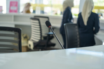 Close-up of a conference microphone on a white desk with blurred female office workers in background, illustrating a modern communication setup in a professional workspace.